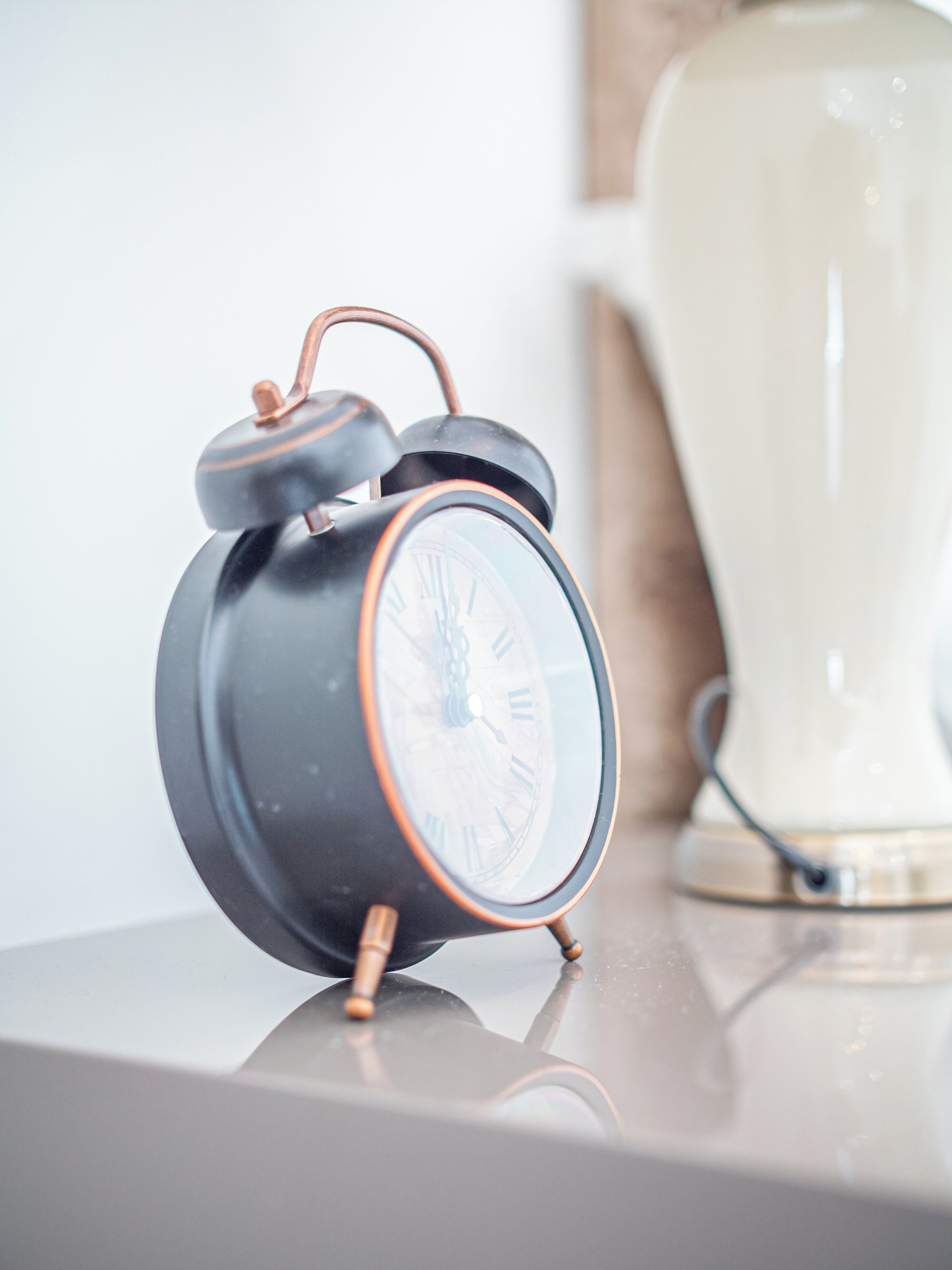 A black alarm clock with Roman numerals on its face, placed on a reflective surface.