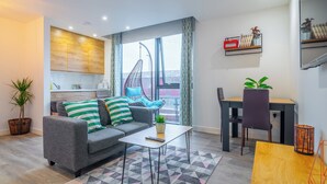 A modern living room with a grey sofa, striped pillows, a hanging chair, a dining table, and a kitchen area with a wooden wall panel. - Priestley Street Apartment in Sheffield (Sheffield)
