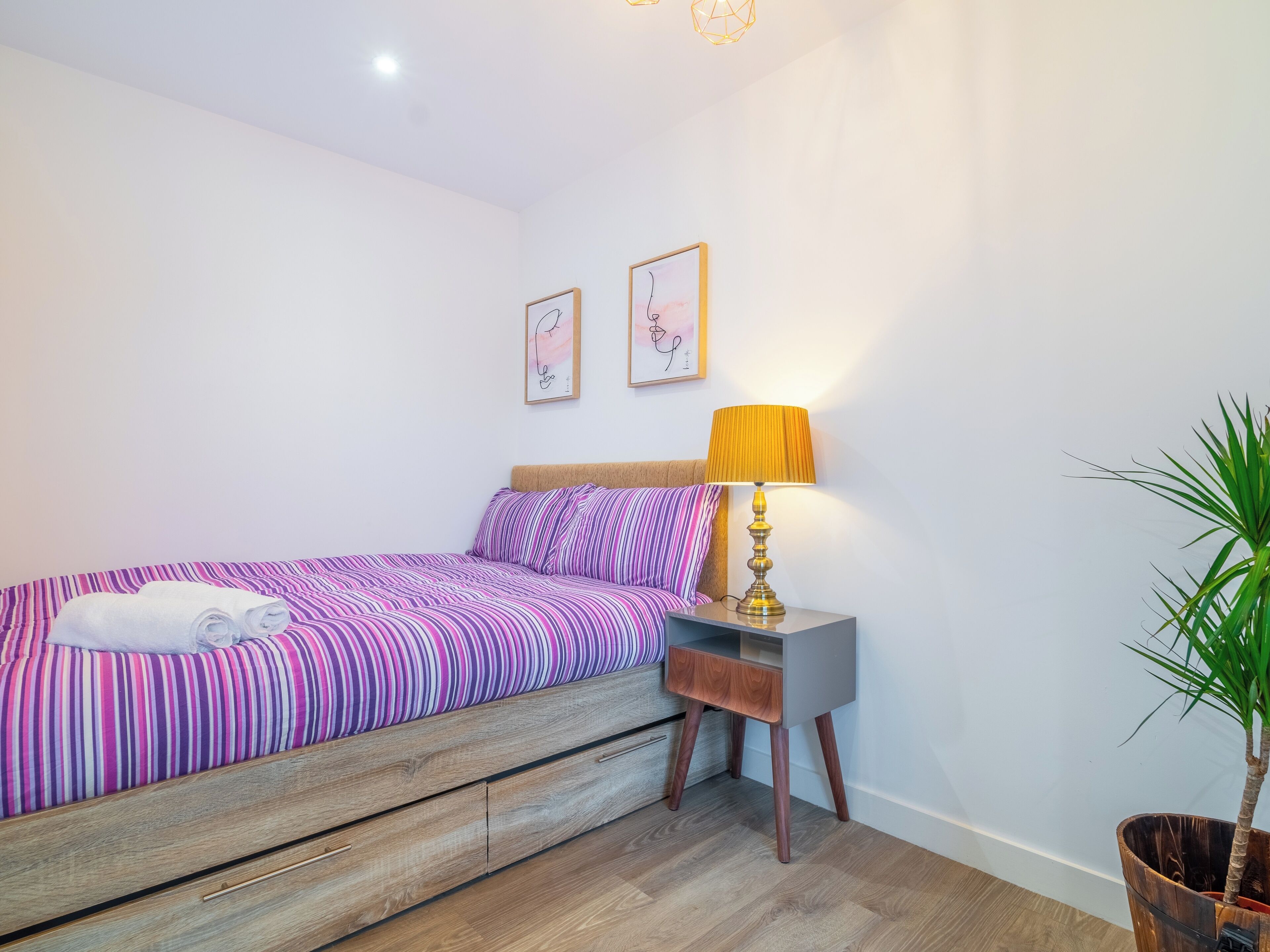 A bedroom with a bed, a nightstand, a lamp, and a potted plant.
