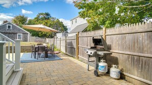 A backyard patio with a grill, a dining table with chairs, and a wooden fence.