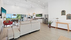 A spacious living area with a sofa, red armchair, and dining table. A kitchen with modern appliances and a large window is visible in the background.