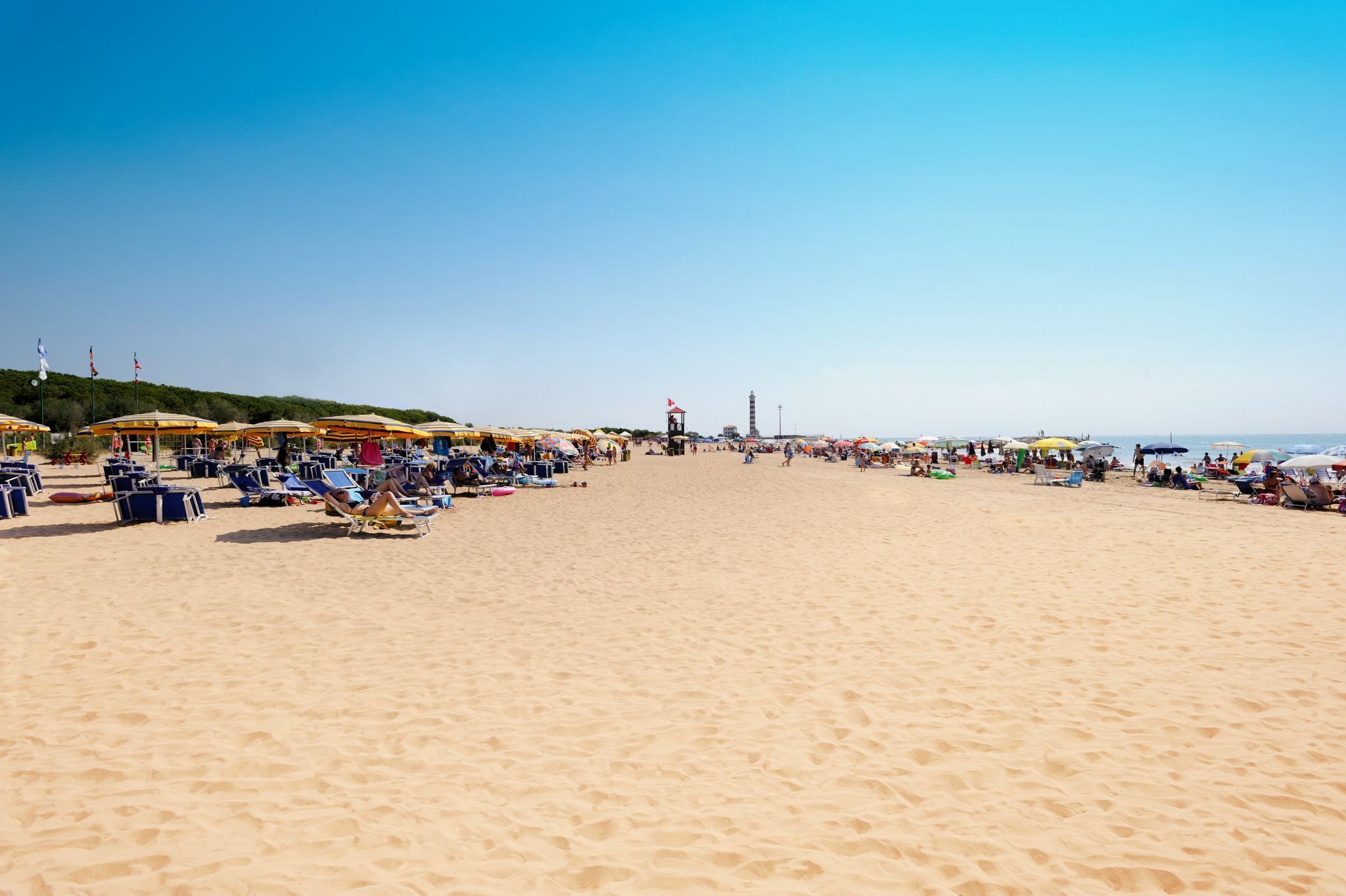 Een breed zandstrand met veel parasols en ligbedden, een redershut en een helderblauwe lucht.