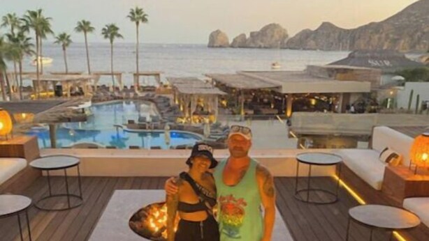 A couple posing on a wooden deck with a pool and lounge chairs in the background.