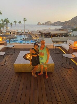 Una pareja posando en una terraza de madera con una piscina y sillones de descanso al fondo.
