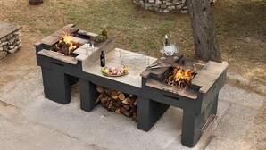 Ein draußen aufgebauter Grillbereich mit zwei Feuerstellen, einem Tisch mit Speisen und Getränken sowie einer Steinsäule im Hintergrund.
