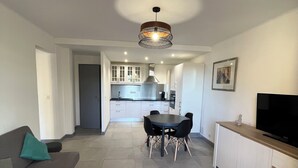 Una sala de estar moderna con un sofá gris, una mesa de comedor con sillas, una cocina al fondo y un televisor sobre un mueble de madera.