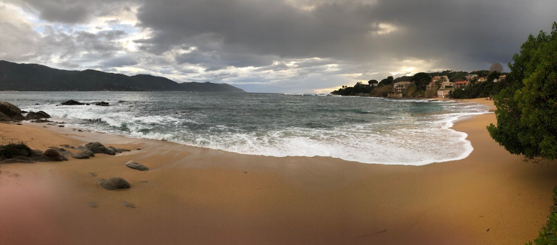 A beach with waves, a mountain in the background, and buildings on the shore.