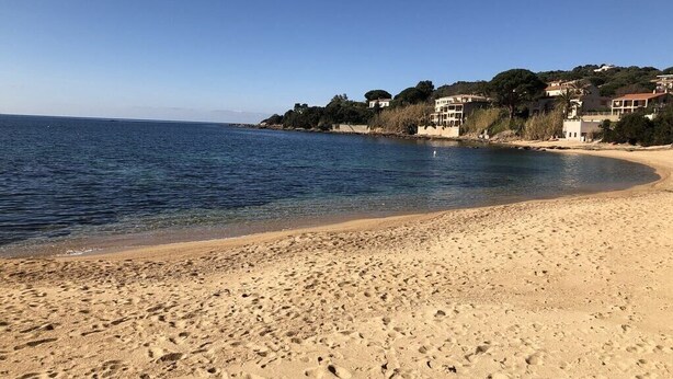 Una playa arenosa con huellas, un mar azul cristalino y edificios al fondo.