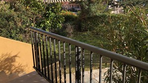Un balcón con toldo a rayas, baranda de metal y vista a un jardín con diversas plantas y un edificio al fondo.
