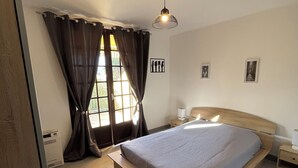 Un dormitorio con cama, mesita de noche, cortinas en la ventana, una lámpara de pared y un pequeño marco de fotos.
