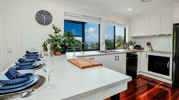 Una cocina moderna con armarios blancos, un ventanal con vistas, un reloj y un comedor con platos azules y copas de vino.
