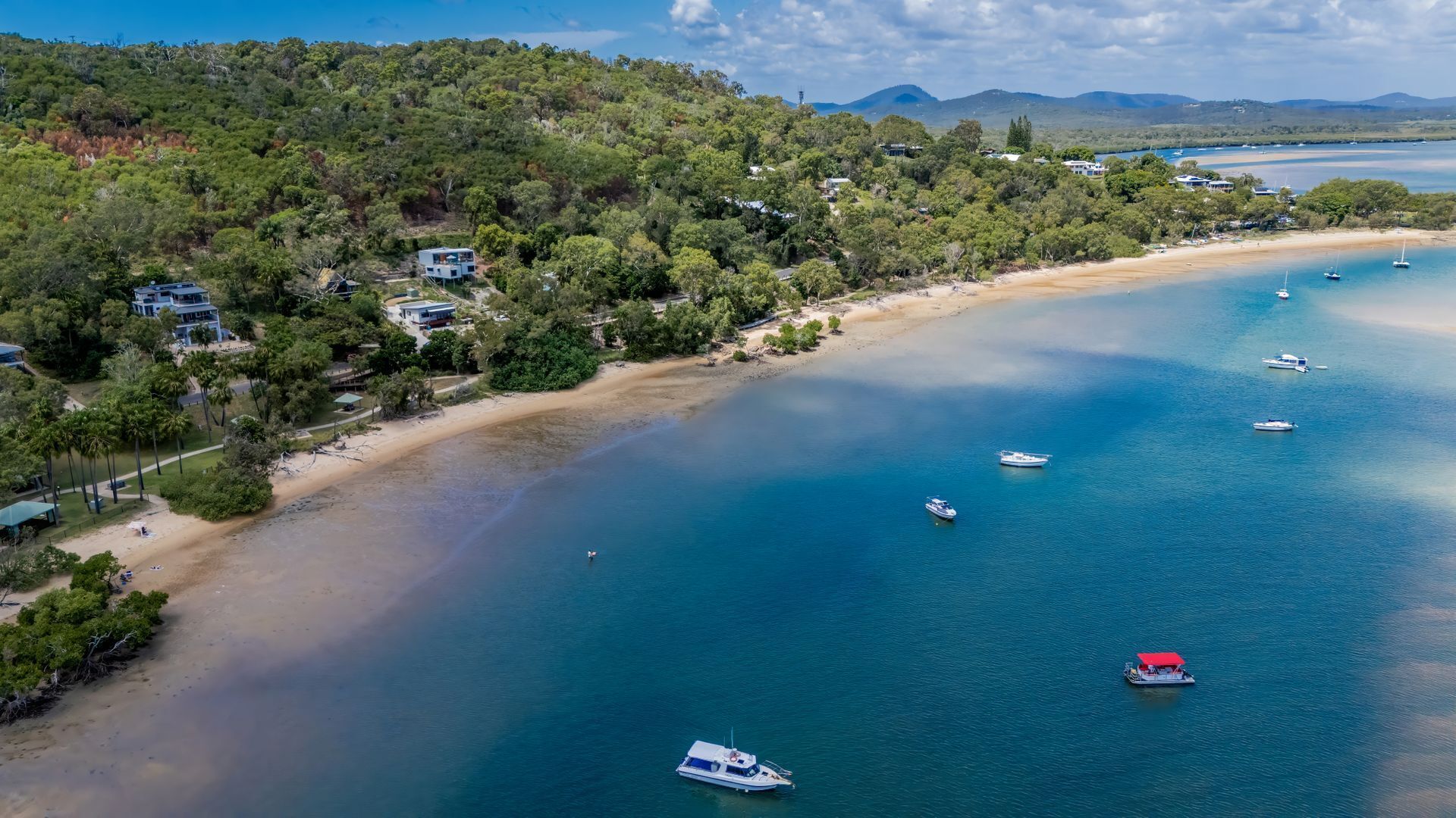 An aerial view of a coastal area with a sandy beach, several boats, and houses surrounded by dense greenery.