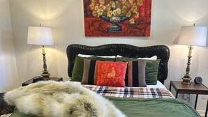 Un letto con una coperta verde e bianca, una soffice coperta in pelliccia e numerosi cuscini. Due comodini con lampade sono presenti ai lati. Sopra il letto, un quadro raffigura dei fiori.