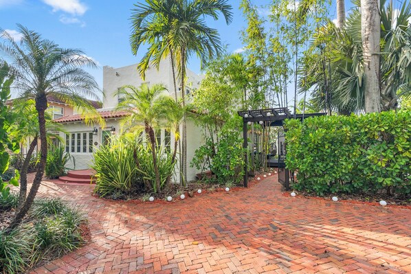 A garden pathway leading to a house with a white facade and red-tiled roof, surrounded by lush greenery and palm trees. - 2 Mansions Pool Garden & BBQ Sleeps 22-8 BR (West Palm Beach)