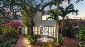 Un jardín con una fuente, palmeras y un edificio blanco con ventanas.