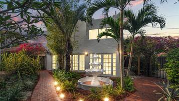 A garden with a fountain, palm trees, and a white building with windows.