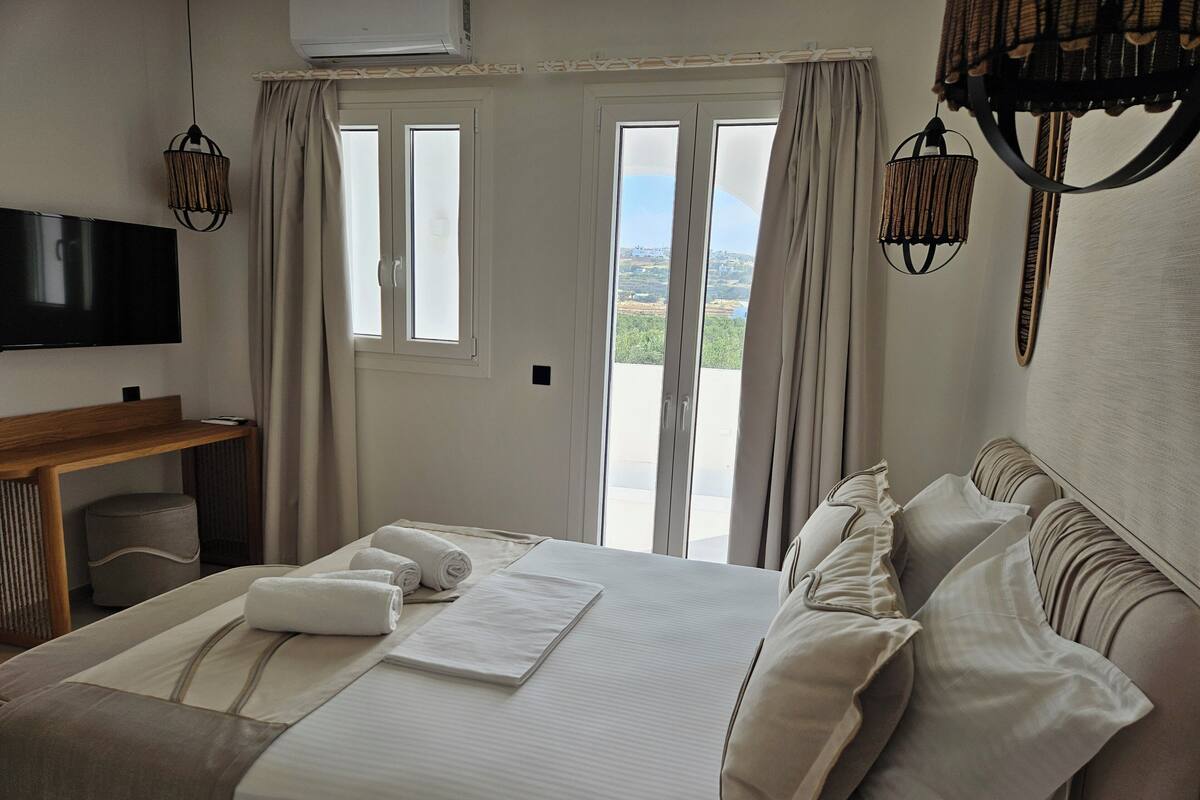 Ein Hotelzimmer mit Bett, Schreibtisch, Flachbildfernseher und Blick ins Freie durch die Fenster.