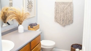 A bathroom with a vanity, a toilet, a hanging macrame wall decoration, and a basket with towels. - Boot Scoot'n In the Stockyards. 3 bedrooms/2 bath (Fort Worth)