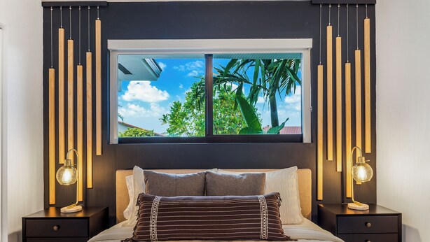 Ein modernes Schlafzimmer mit großem Fenster, zwei Nachttischen mit Lampen, einem Bett mit gestreifter Bettwäsche und Blick auf Palmen.