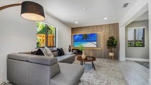 Ein modernes Wohnzimmer mit einem grauen Sofa, einem Couchtisch und einem großen Fernseher, der eine Strandlandschaft zeigt.