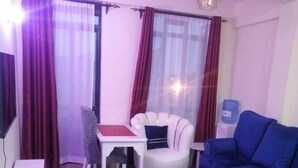 Sala de estar con un sofá blanco, un sofá azul, una mesita blanca y un tapiz rojo. Hay una ventana con cortinas y una lámpara colgante.