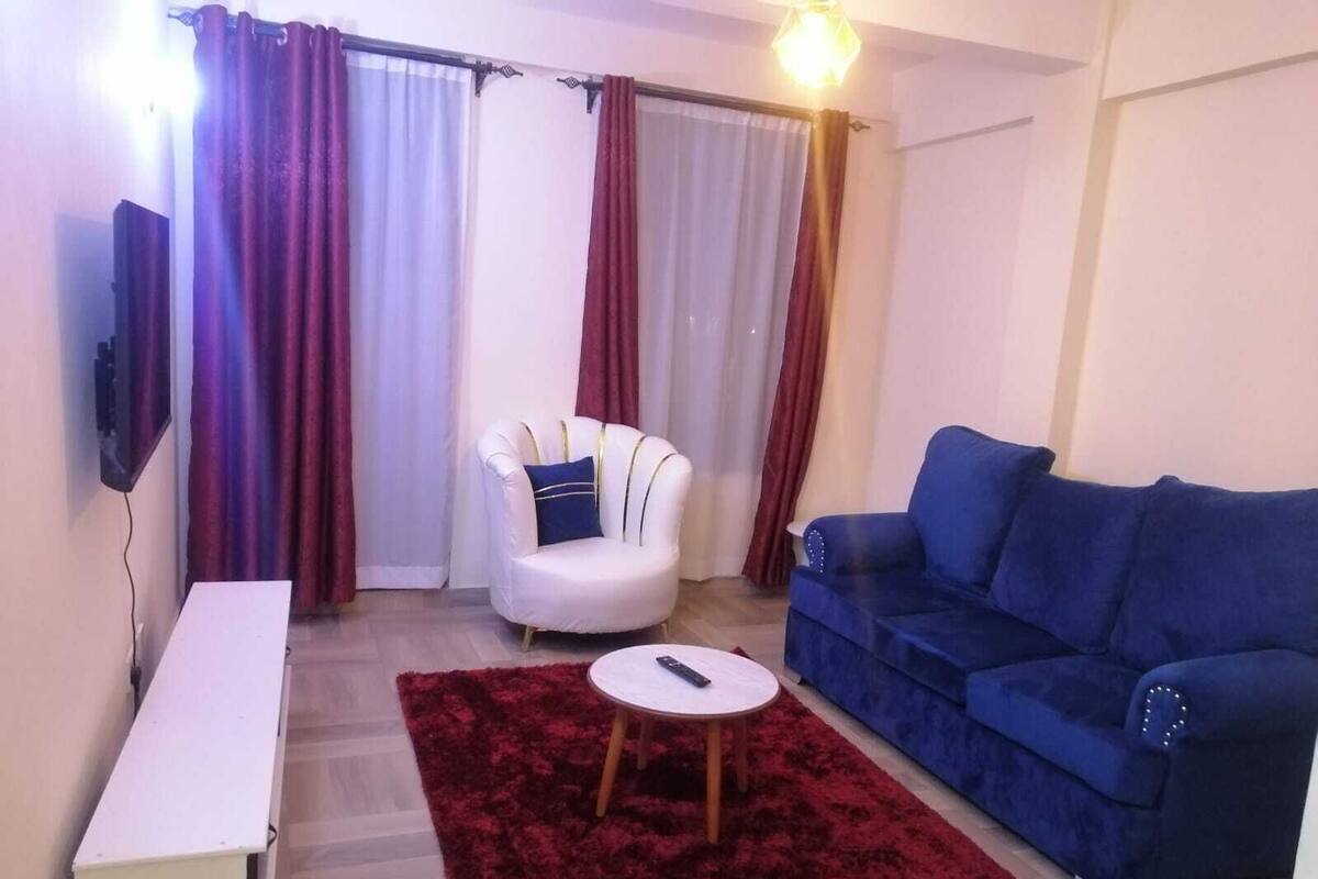 A living room with a white armchair, a blue sofa, a red rug, and a TV mounted on the wall.