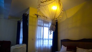 Una habitación de hotel con cama, una silla, ventana con cortinas y una lámpara colgante.