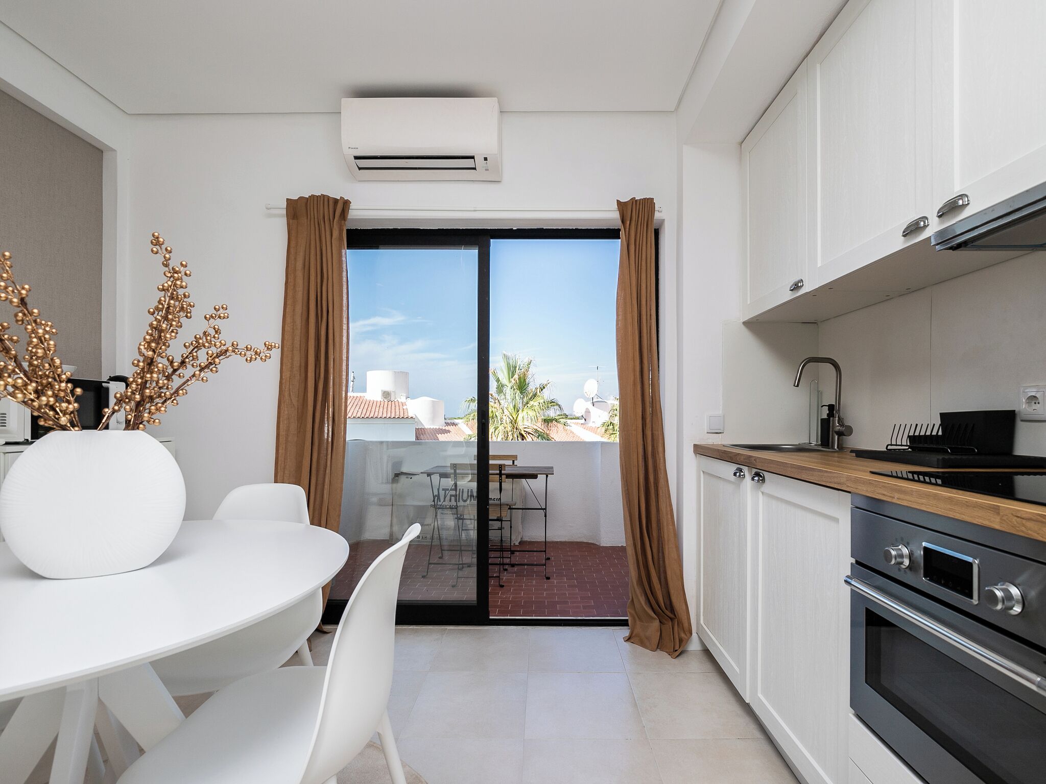 A modern kitchen with a dining area, a sliding glass door to a balcony, and a view of the cityscape.