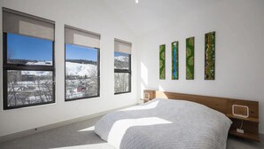 Une chambre avec un grand lit, trois fenêtres donnant sur un paysage enneigé et trois tableaux verticaux accrochés au mur.