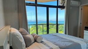 Een slaapkamer met een groot raam met uitzicht op zee en groen.