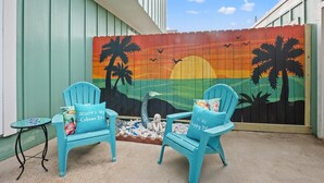 Un patio con dos sillas turquesas, una mesa redonda pequeña y un mural que muestra un atardecer con palmeras y aves.