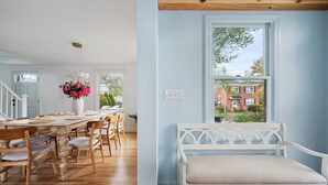 Un comedor con mesa y sillas de madera, un banco blanco y una ventana con vista a un edificio de ladrillo.