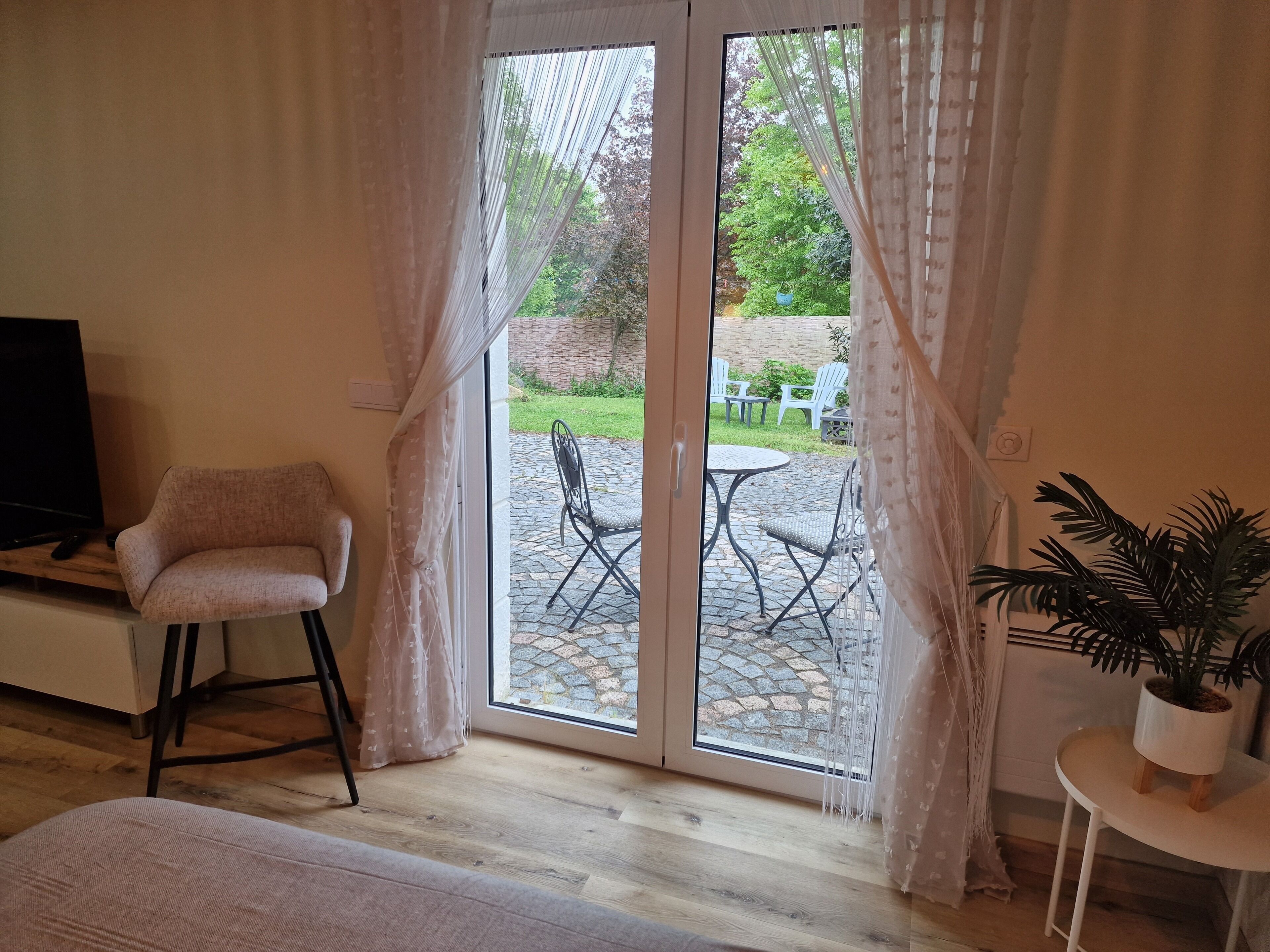 Ein Zimmer mit Fernseher, einem gepolsterten Sessel, einem kleinen Tisch mit Pflanze und einer Schiebeglastür zu einer Außenpatio mit Stühlen und Tisch.