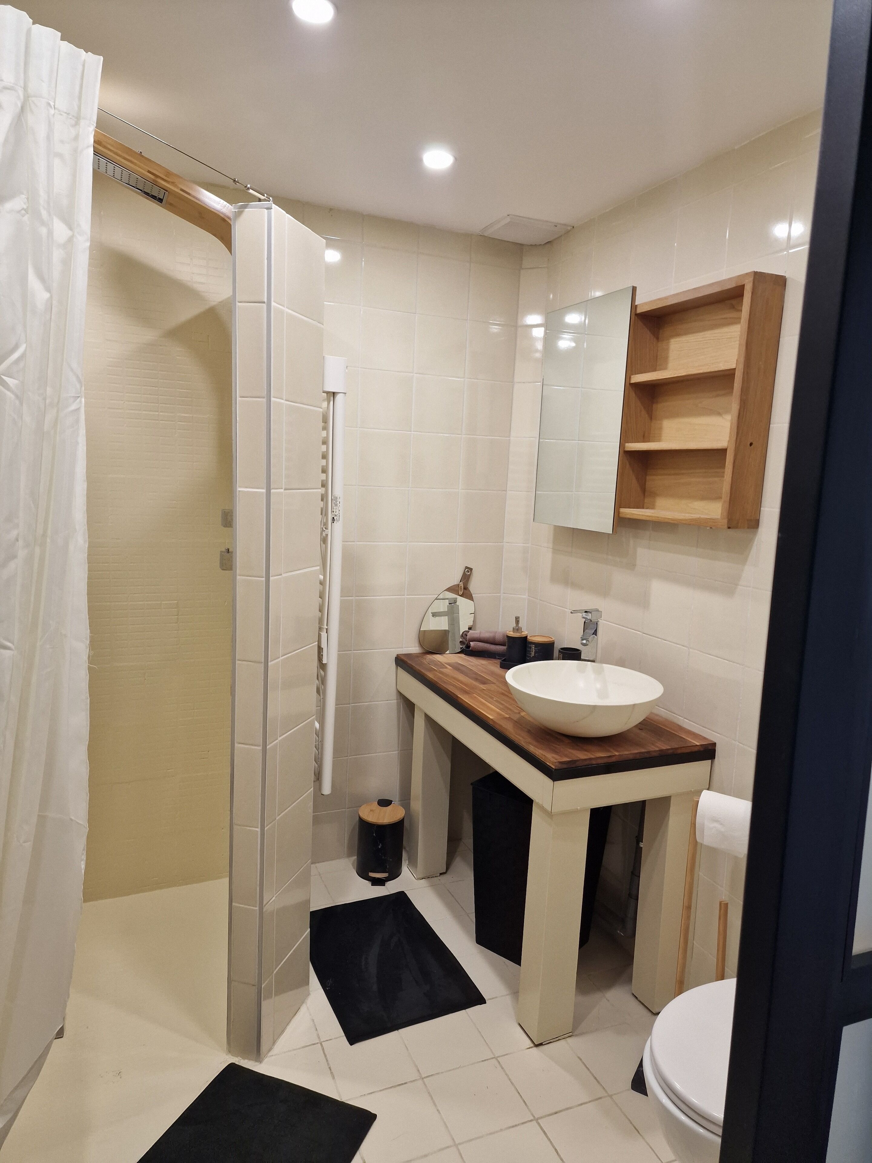 Une salle de bain moderne avec un meuble-lavabo en bois, un lavabo blanc et un miroir au-dessus. Il y a un espace douche avec une porte en verre et un rideau blanc.