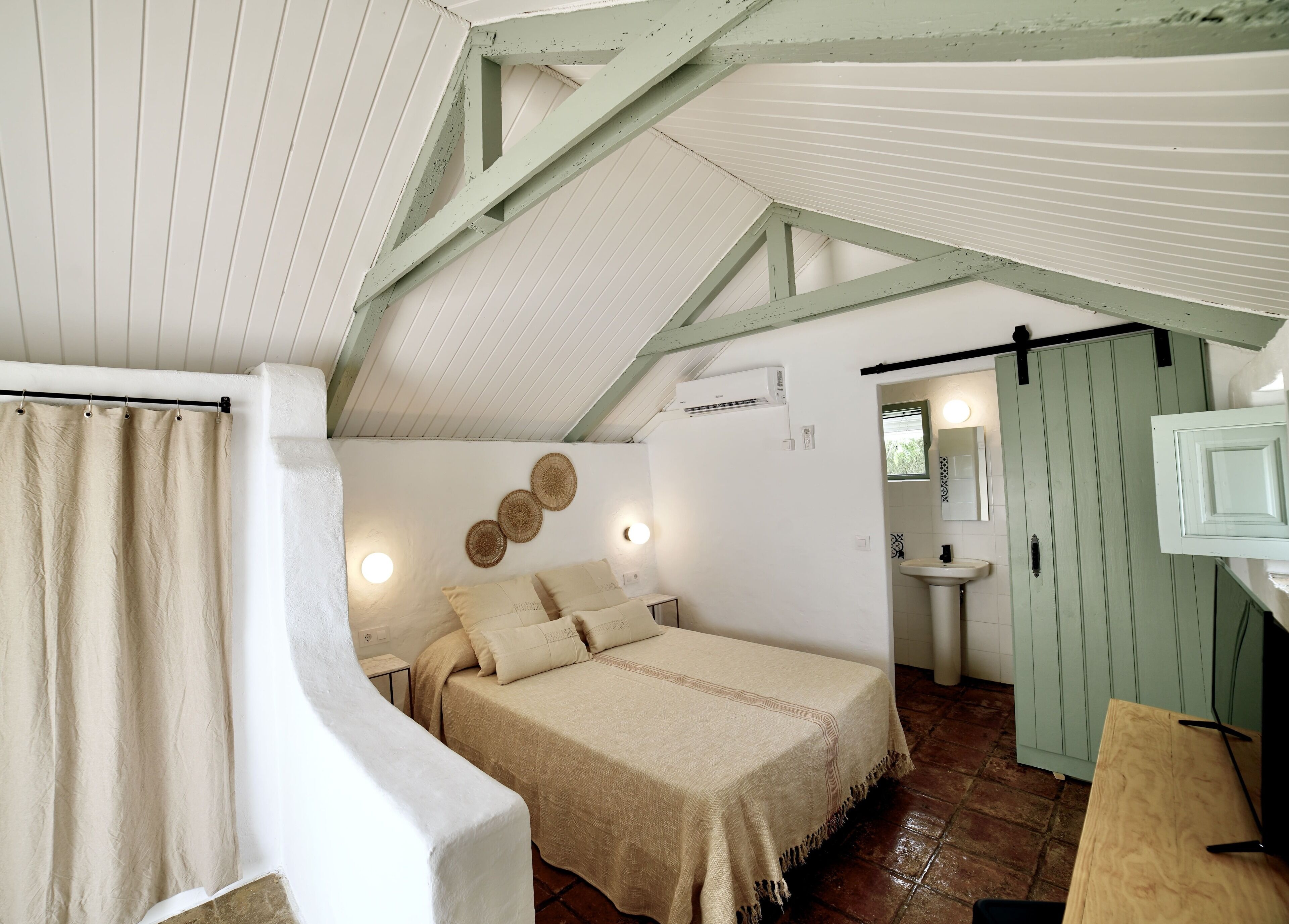 A bedroom with a bed, a bedside table, a mirror, a sink, and a window.