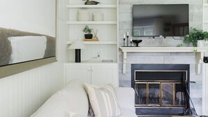 A modern living room with a white sofa, a fireplace, and a mounted TV. - Le Pavillon | Comfort in Nature (Saint-Côme)