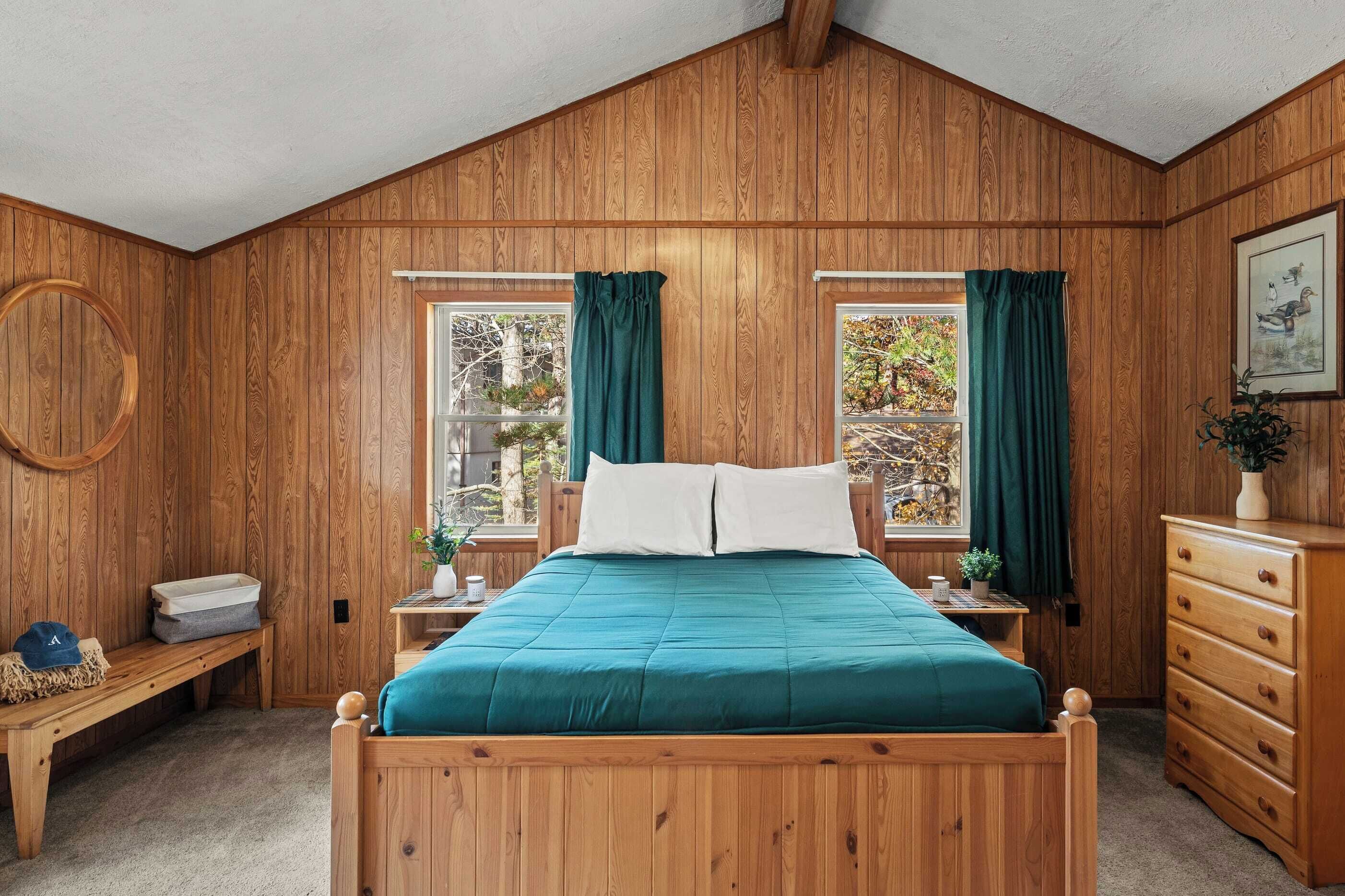 Une chambre à coucher aux murs lambrissés, avec un lit, deux fenêtres avec des rideaux, une commode en bois, un banc et un miroir rond.