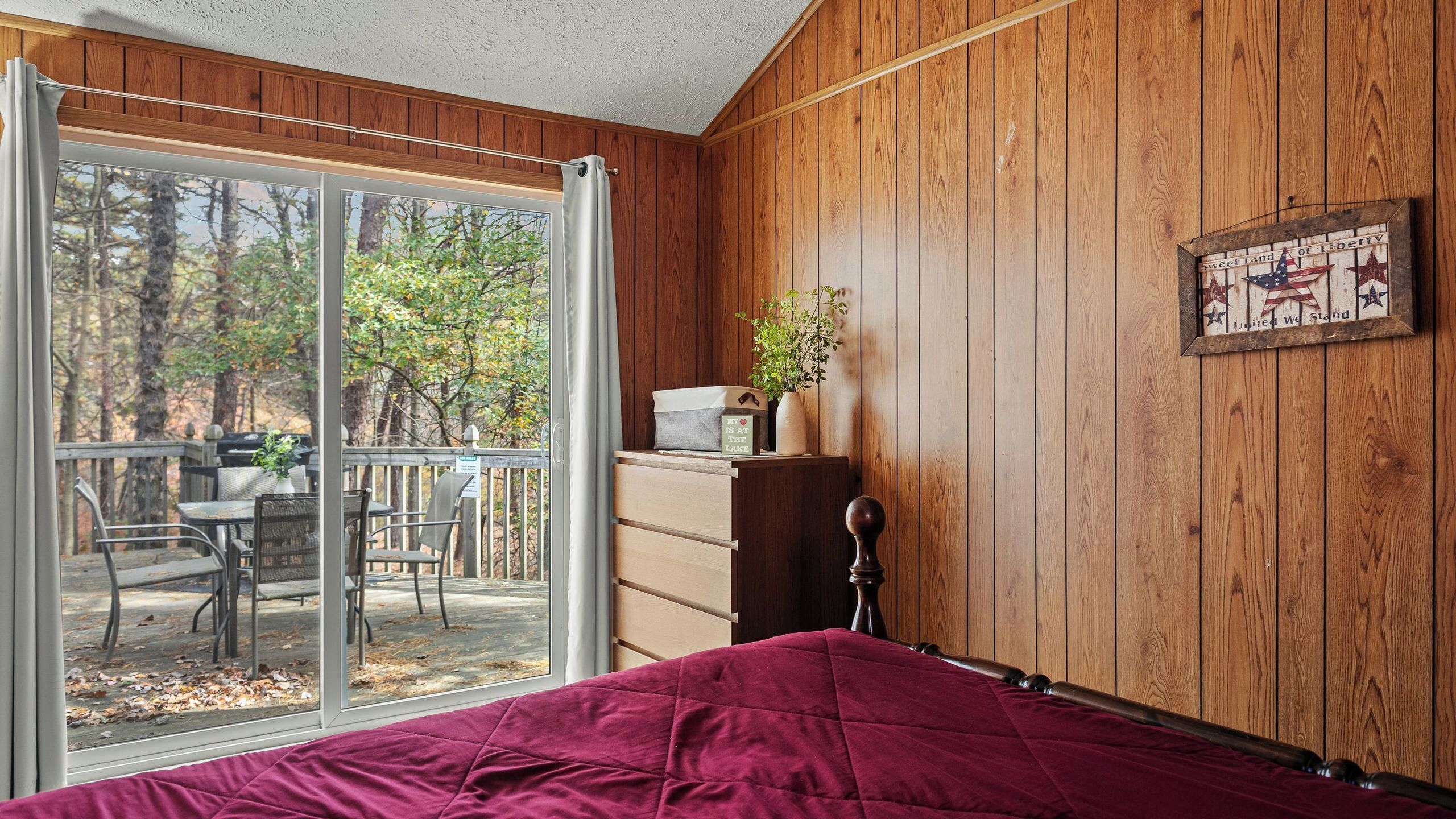 A room with a bed, a wooden dresser, a potted plant, a framed sign, and a sliding glass door leading to an outdoor deck.