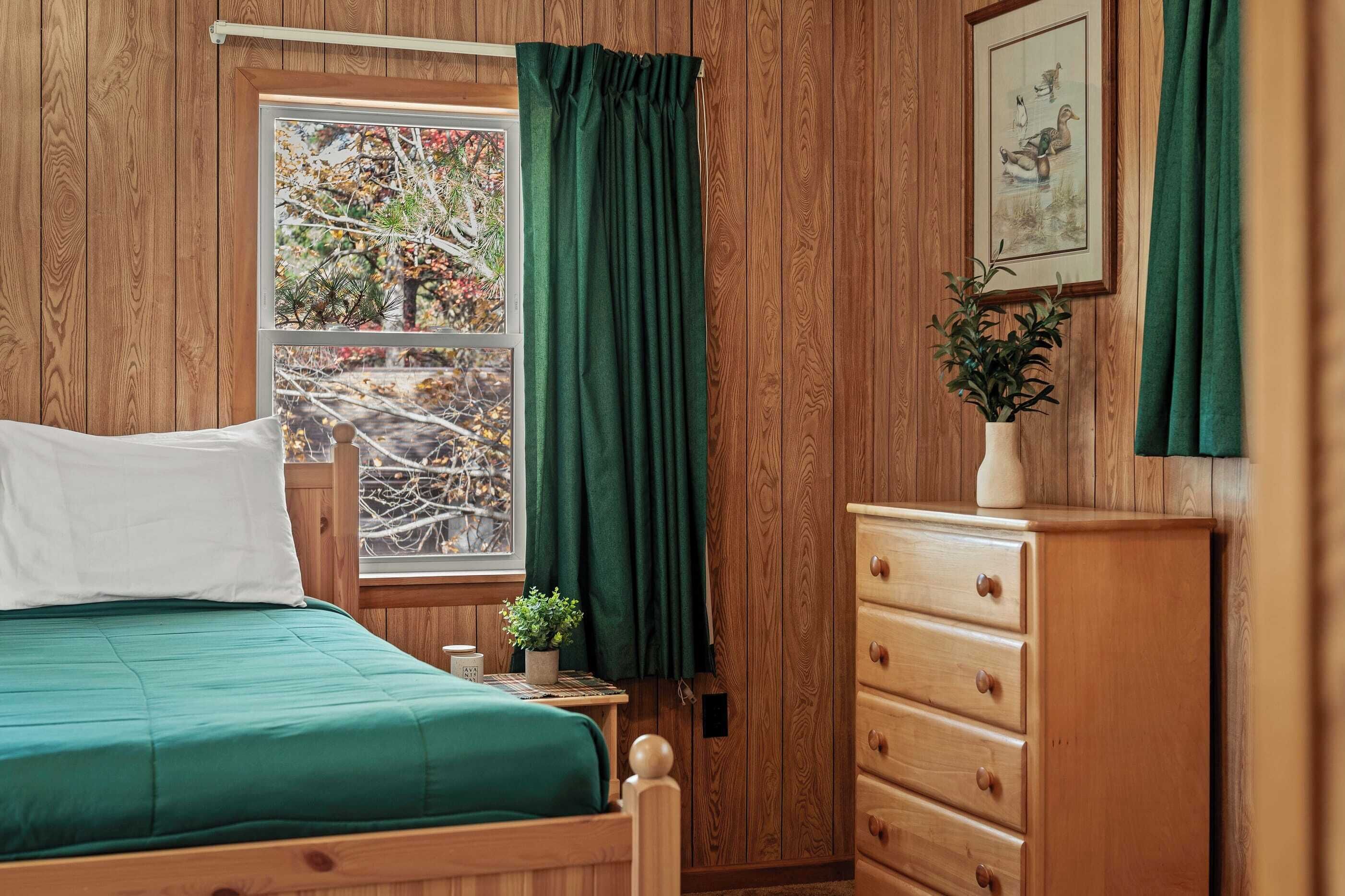 Une pièce aux murs lambrissés, comprenant un lit, une commode, une fenêtre avec des rideaux et un tableau encadré accroché au mur.