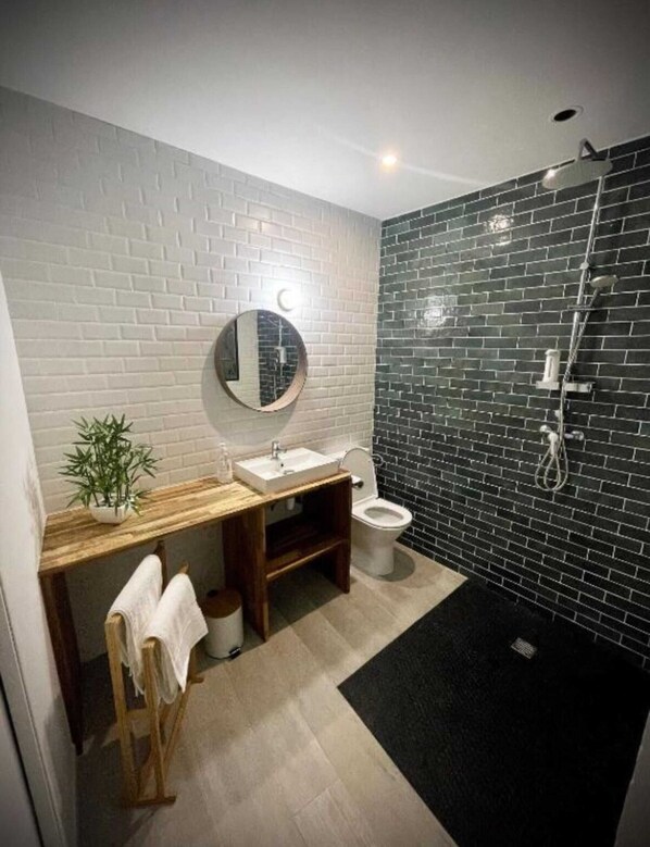 A bathroom with a white brick wall, a wooden vanity, a round mirror, a toilet, and a shower area with black tiles.