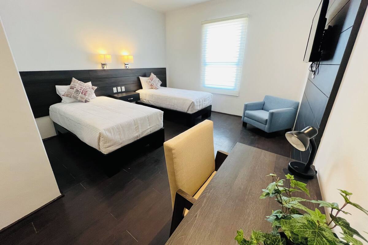 Une chambre d’hôtel avec deux lits, un bureau, une chaise, une télévision et une fenêtre avec des stores.