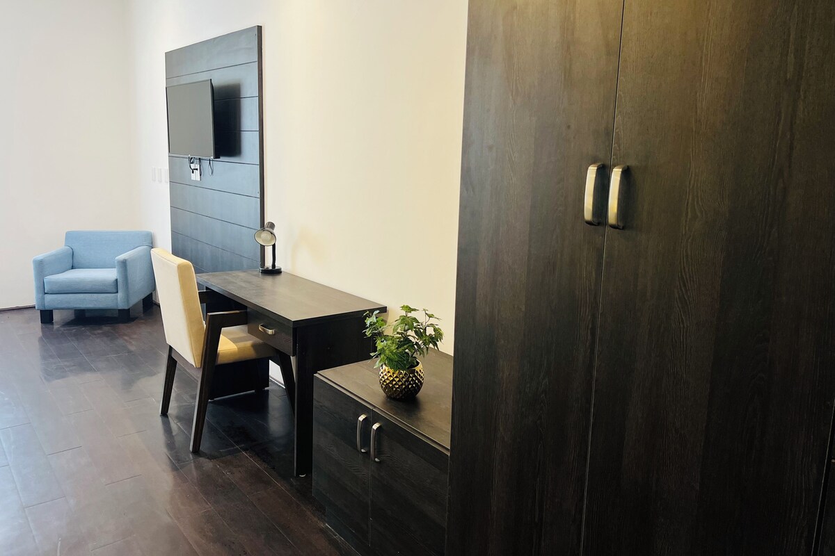 Quarto moderno com escrivaninha de madeira escura, cadeira azul clara, espelho grande e uma pequena planta em vaso.