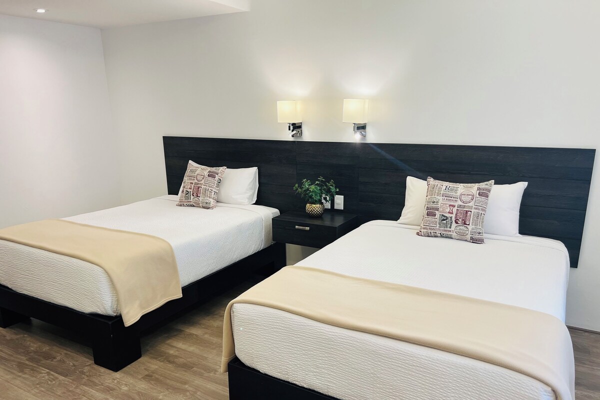 A hotel room with two beds, each with a white bedspread and a beige throw blanket. There are two wall-mounted lamps above the beds, and a small table with a plant between them.