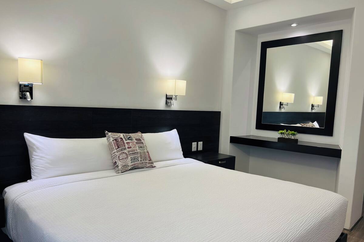 Une chambre d’hôtel avec un grand lit, une tête de lit noire, deux lampes de chevet, un miroir fixé au mur et une petite étagère avec des plantes.