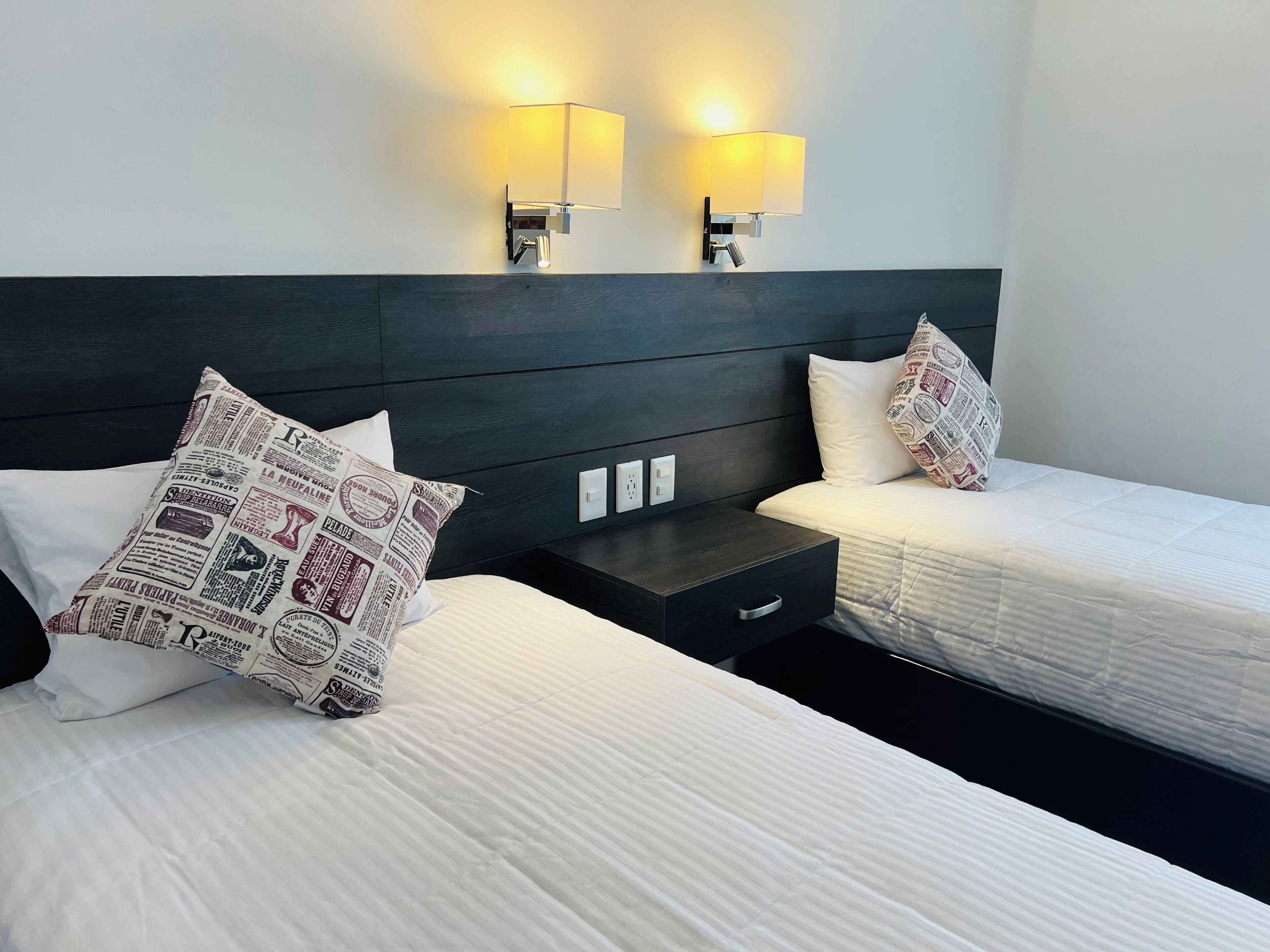 Habitación de hotel con dos camas, cada una con una almohada y una mesita de noche. Dos lámparas fijadas a la pared proporcionan iluminación.