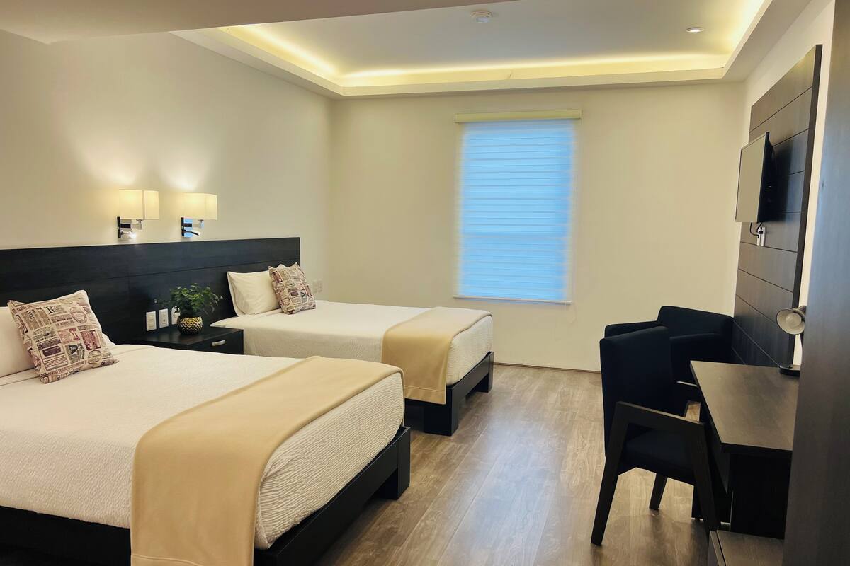 Une chambre d’hôtel avec deux lits, un bureau, une chaise, une télévision et une fenêtre avec des stores.