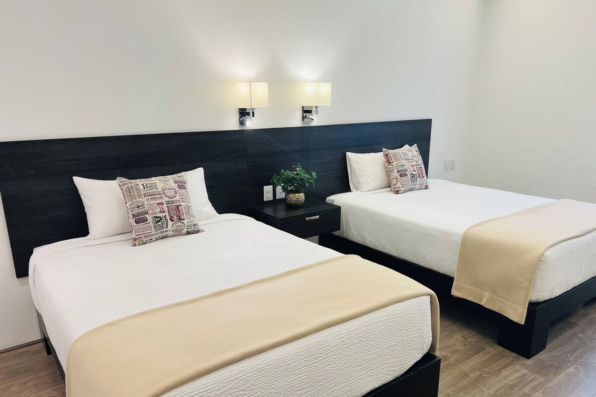 Deux lits avec du linge de lit blanc et des plaids beiges, une tête de lit sombre et deux lampes fixées au mur.