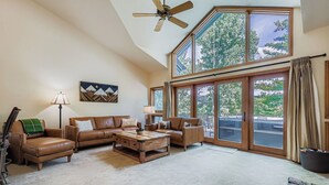 A spacious living room with a high ceiling, a ceiling fan, a large window with a view of trees, a brown leather sofa, a coffee table, and a lamp.
