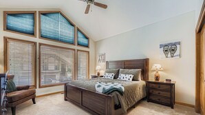 A bedroom with a large bed, wooden headboard, multiple windows with blinds, a ceiling fan, a nightstand with a lamp, and a chair.
