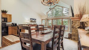 A spacious kitchen with wooden cabinets, a dining area with a large wooden table and chairs, a stone fireplace, and a view of the outdoors through large windows.